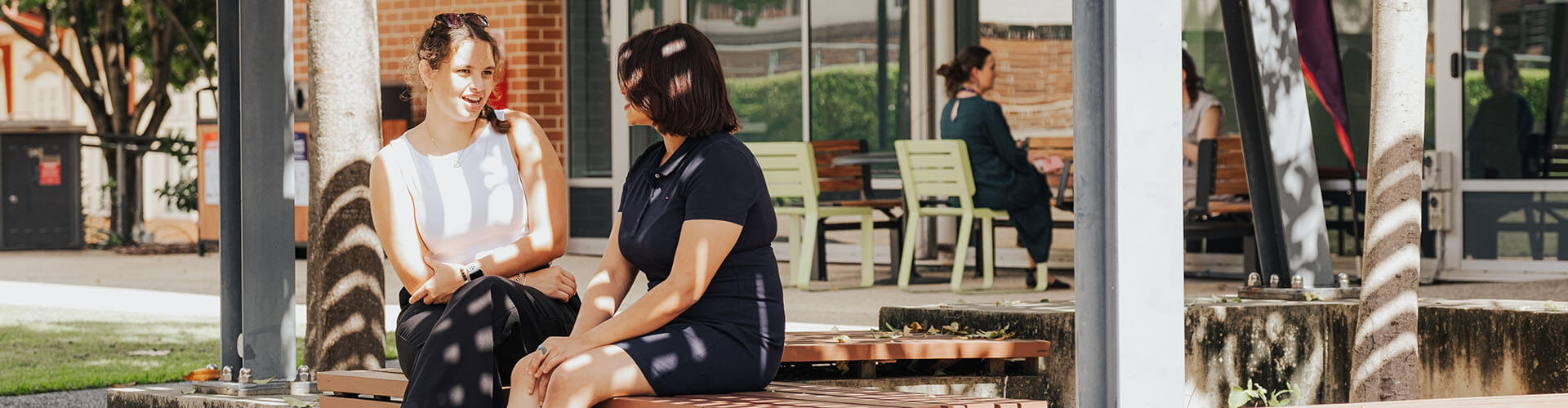 Two women sit on a bench outdoors having a conversation, with a building and other people in the background.