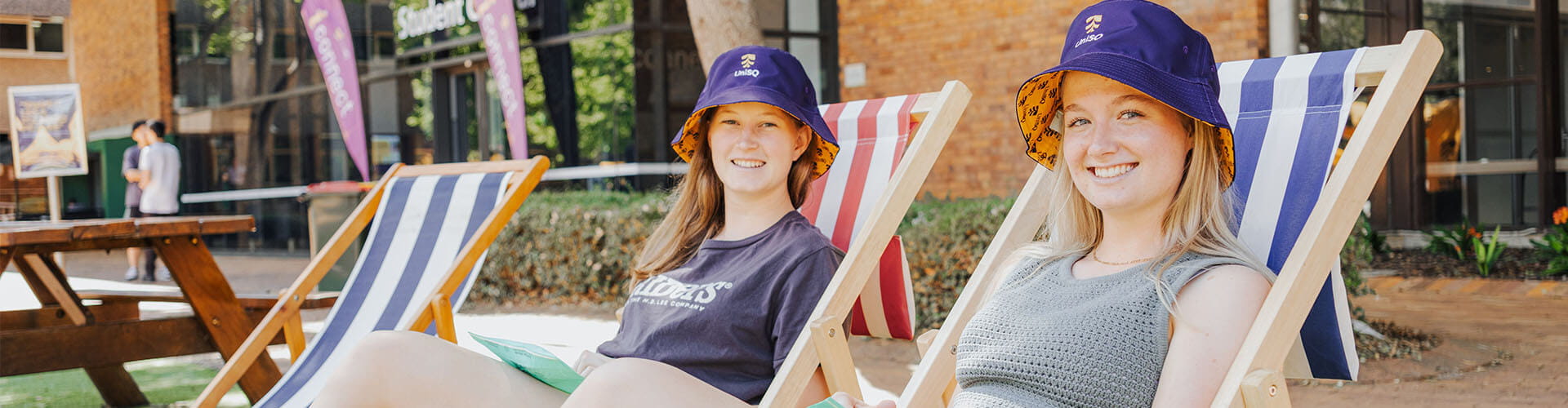 Two young women sit on striped deck chairs outdoors, smiling at the camera and wearing blue bucket hats with orange trim.