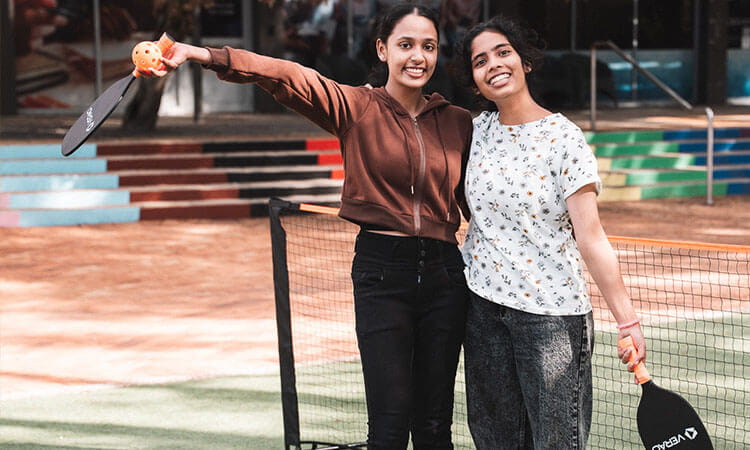 Two young women stand on a pickleball court, smiling at the camera, each holding a pickleball paddle and one holding a ball.
