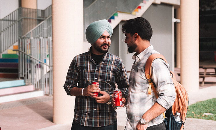 Two men stand outside talking and holding cans of Coca-Cola; one has a backpack and the other is wearing a turban.