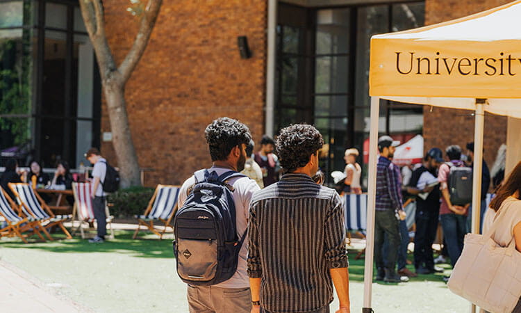 Students with backpacks walk past a university event tent outdoors, with other people sitting on deck chairs and gathering nearby.