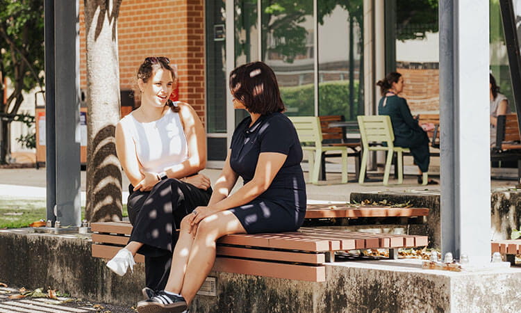 Two people sit on a wooden bench outdoors, engaged in conversation. Other individuals are seated in the background near tables and trees.