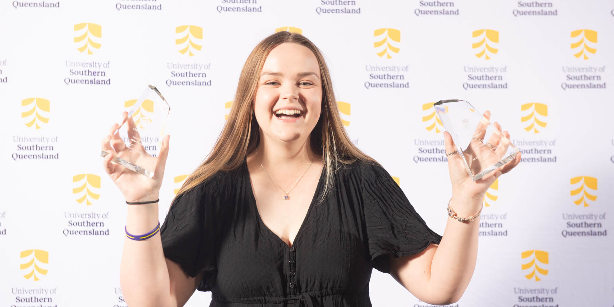 A person smiles while holding two awards, standing in front of a backdrop with the University of Southern Queensland logo.