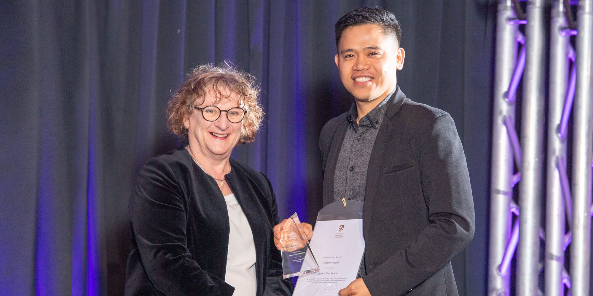 Two people stand indoors; one, a woman with curly hair and glasses, hands an award to a smiling man dressed in a dark blazer. They are posing for a photo against a backdrop of dark curtains and vertical lights.