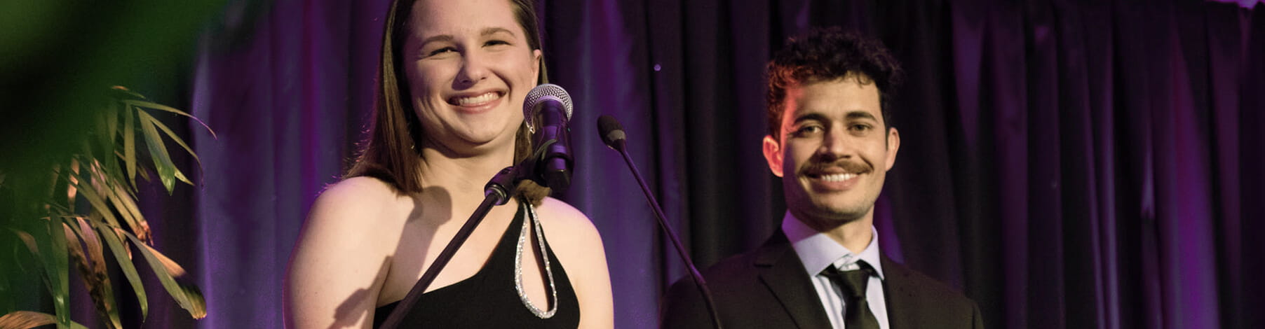 A woman in a black dress laughing beside a man in a suit, both standing on stage with a purple curtain backdrop.