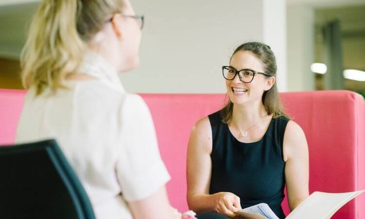 Two women engaging in a conversation while sitting on a pink sofa, one holding a document.