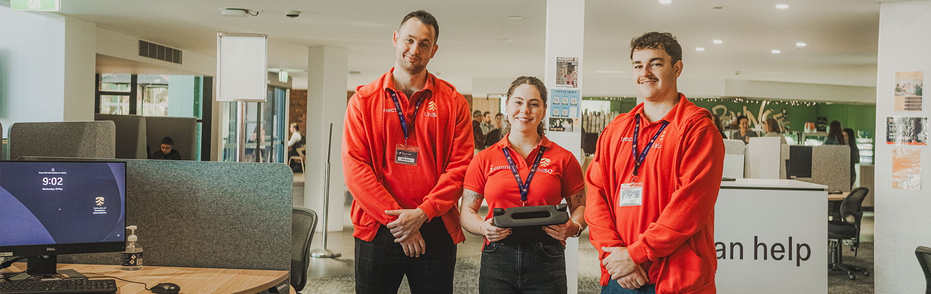 Three people wearing red shirts and badges stand together in an office space with various workstations and a "can help" sign in the background.