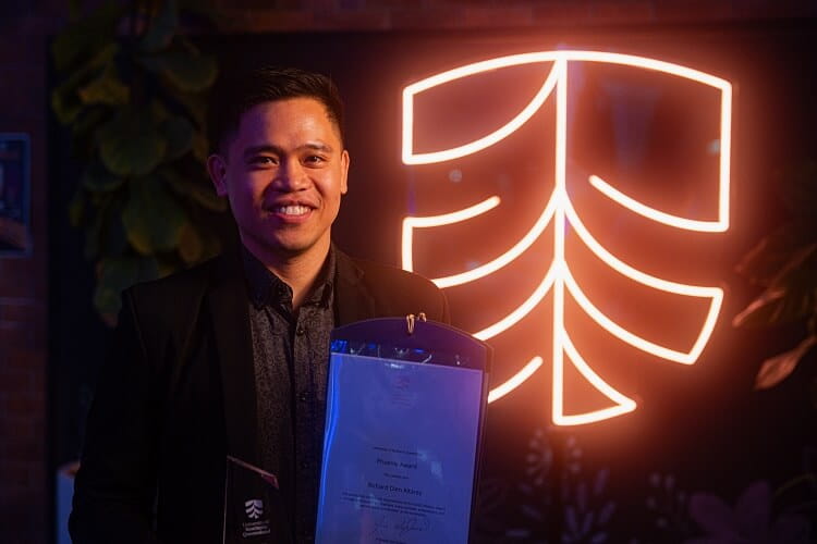 A person stands smiling, holding an award and a certificate, with a glowing emblem in the background.