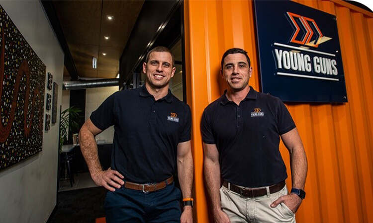 Two men in business casual attire posing in front of a sign that reads "young guns" in an office setting.