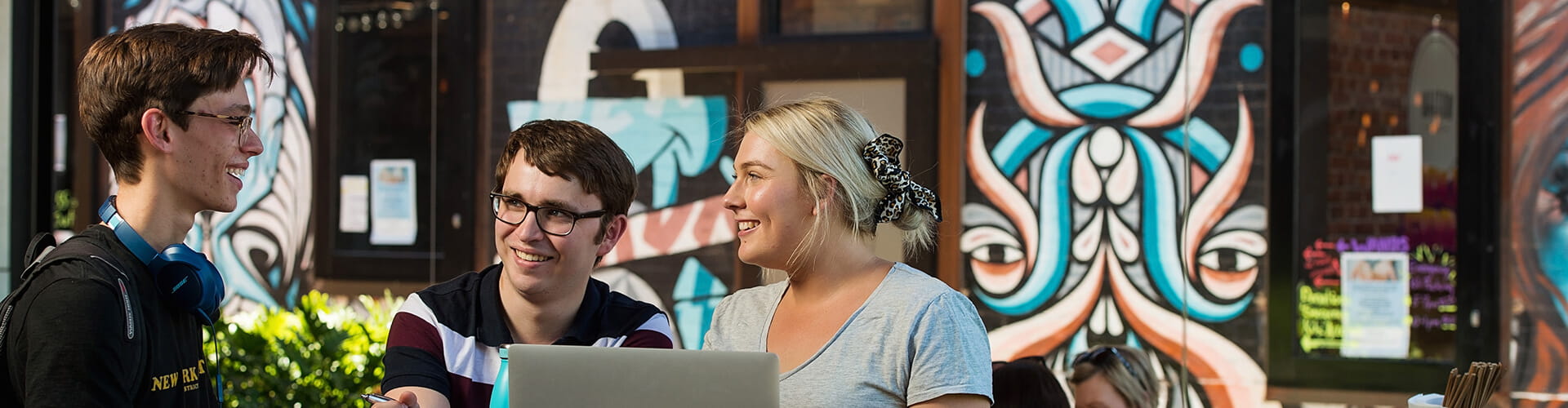 Three young adults engaging in a cheerful conversation, with one holding a laptop, against a backdrop featuring urban street art.