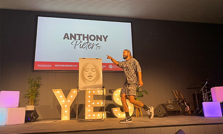 A speaker presenting on stage next to a screen displaying the name "anthony pieters" with artwork that includes the word "yes.