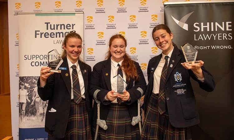 Three young women holding awards in front of a banner.