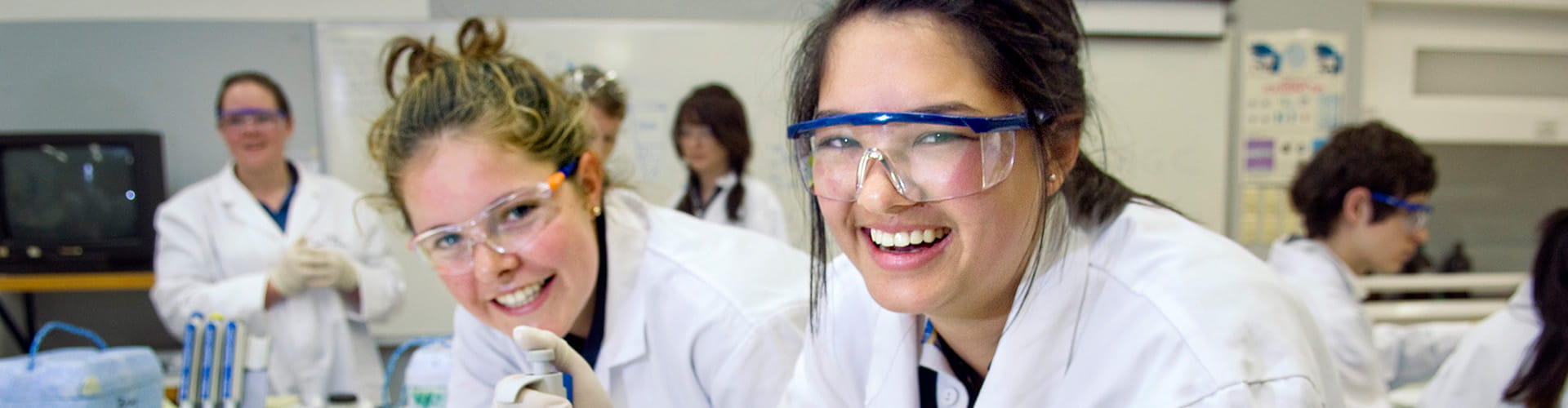 Two women in lab coats and safety glasses smiling in a science laboratory setting.