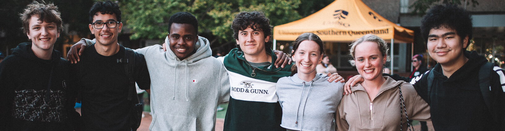 A group of seven young adults standing closely together outdoors, smiling at the camera. A yellow tent and some greenery are visible in the background.