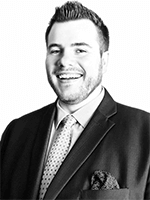 A man with short hair and a beard is wearing a suit with a pocket square and a tie. He is smiling and looking at the camera. The background is plain white.