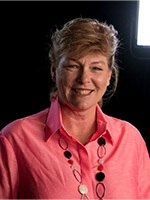 A woman with light brown hair wearing a bright pink shirt and a necklace featuring round beads, smiles in a dimly lit setting.