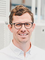 A man with short, curly brown hair and glasses, wearing a white button-down shirt, is smiling in an indoor setting.