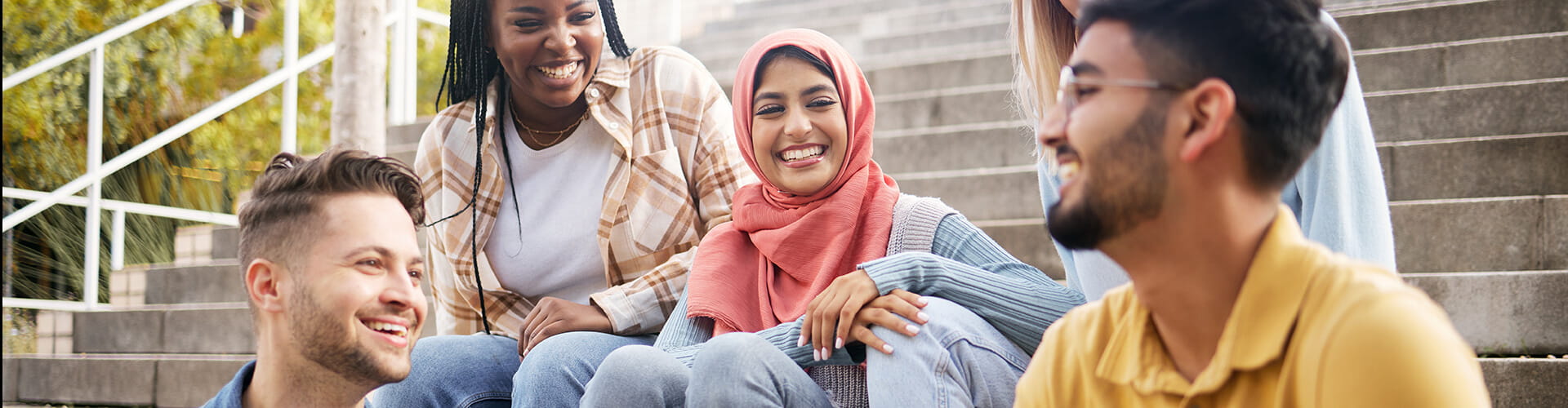 A diverse group of young adults sit on outdoor steps, smiling and engaging in conversation.