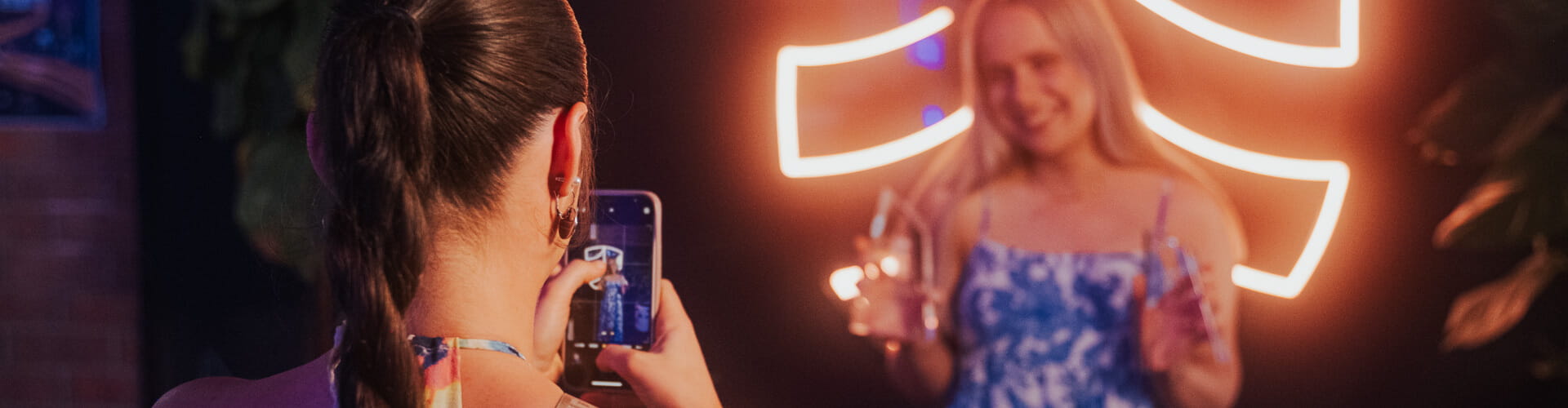 A person with braided hair is taking a photo of another person posing in front of a neon light backdrop, both in a dimly lit environment.