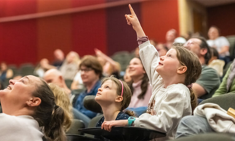Children and adults sit in a theater audience, with a young girl pointing upwards.