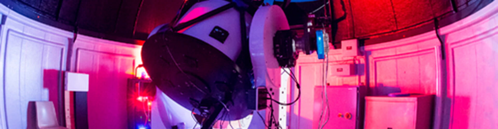 A large telescope in an observatory room with colorful lighting. Various cables and equipment surround the telescope.