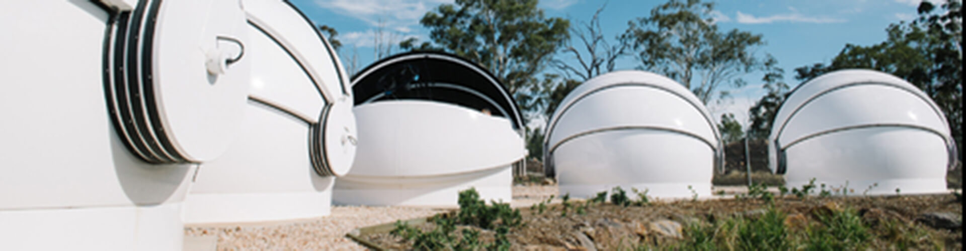 Series of modern, white dome-shaped observatories under a clear sky, surrounded by trees and vegetation.
