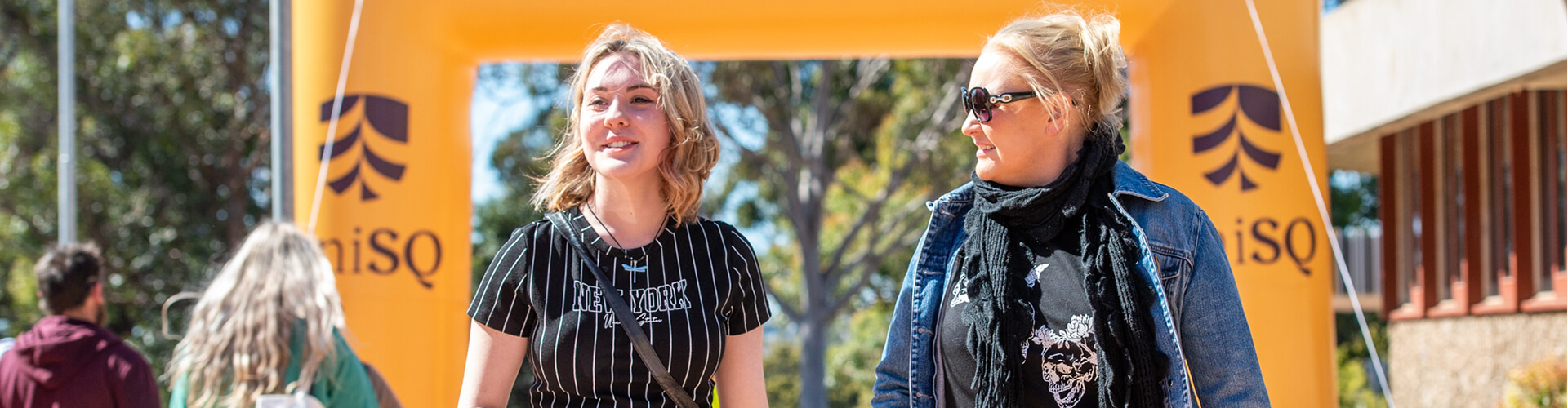 Two women walking outdoors, smiling and talking, with a yellow archway and trees in the background.