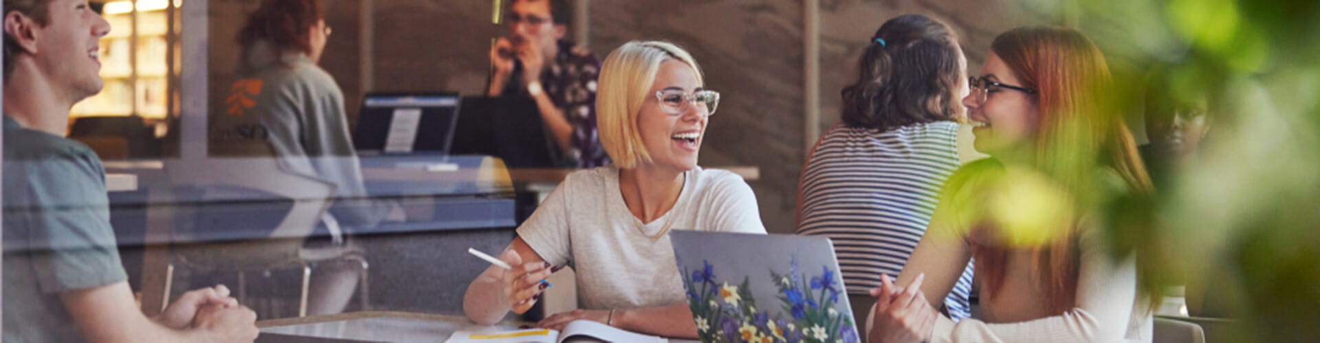 A group of people sitting at a table with laptops and notebooks, engaged in conversation and smiling. A small plant is visible in the foreground.