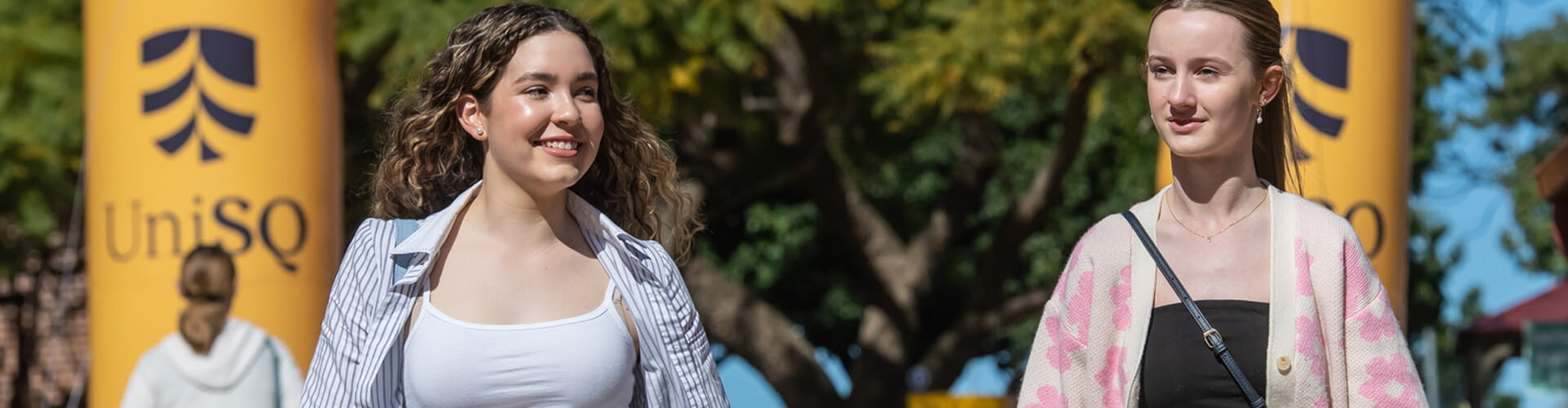 Two people walking outdoors with trees in the background and "UniSQ" banners visible.