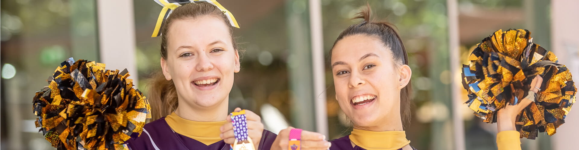 Two cheerleaders in purple and yellow uniforms smile and hold pom-poms outdoors.