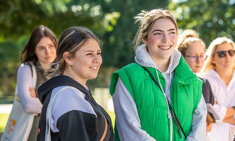 A group of people standing outdoors, with two individuals in focus, one wearing a green vest and the other a black hoodie, smiling while listening.