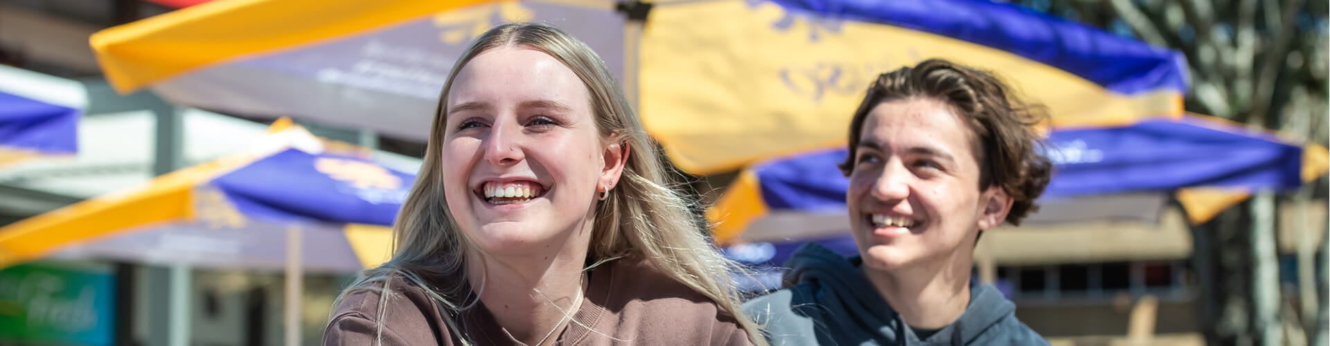 Two people smiling outdoors, sitting in front of yellow and purple umbrellas.