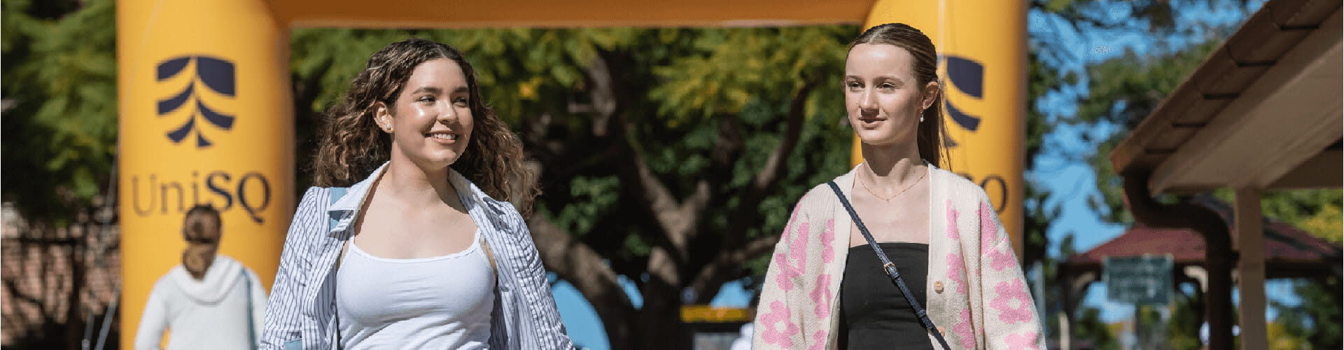 Two women are walking outside, smiling, with trees and a yellow UniSQ arch in the background.