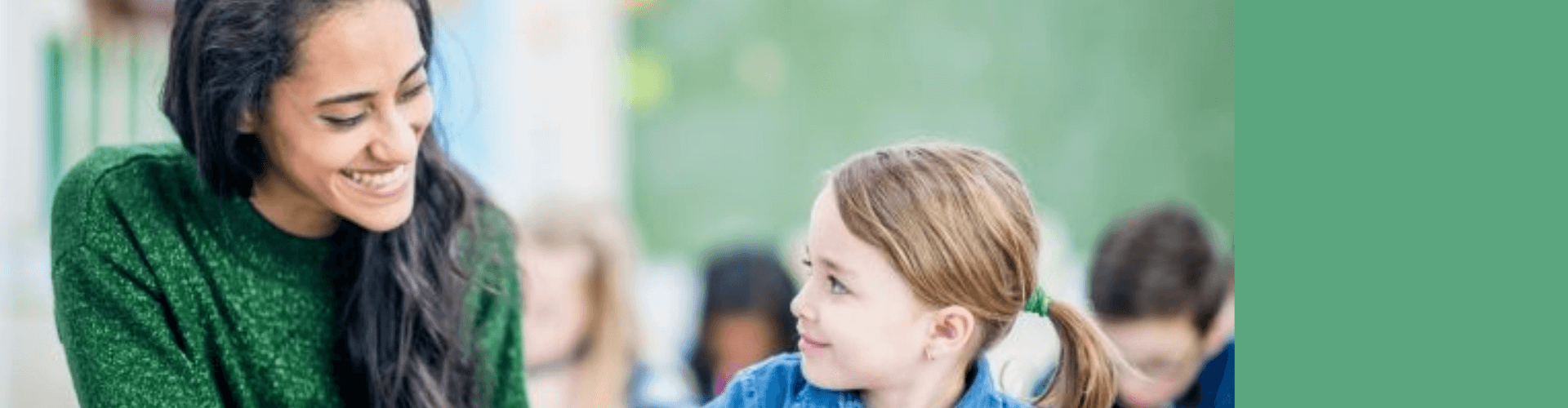 A woman in a green sweater smiles at a young girl with a ponytail. They are sitting together in what appears to be a classroom.