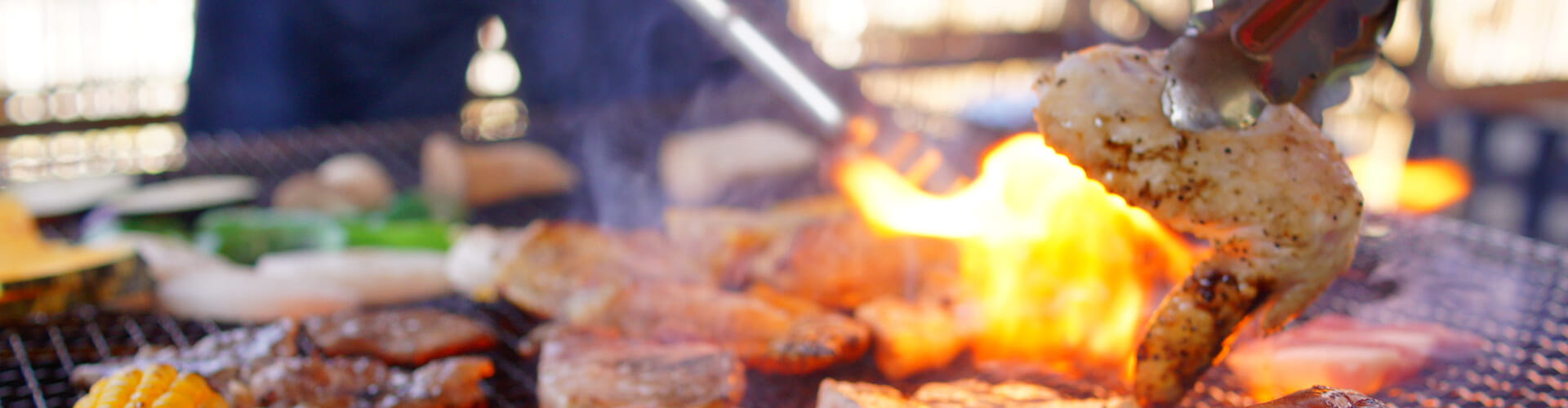 Close-up of a barbecue grill with meat being flipped by tongs over open flames, surrounded by various grilled vegetables.