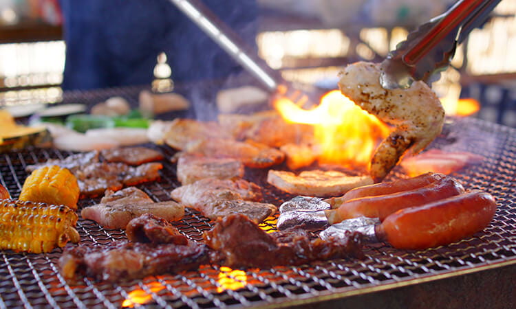 Various meats and vegetables grill on a barbecue with flames visible.