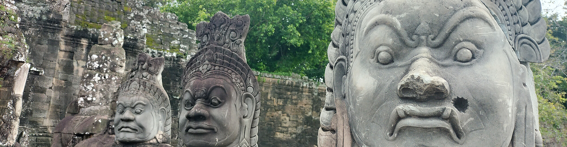 Three stone statues with intricately carved faces stand in a row at Angkor Thom, Cambodia, against a backdrop of ancient ruins and lush greenery.