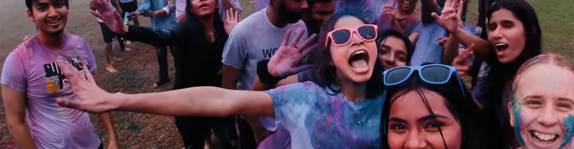 Group of people celebrating with colorful powder, smiling and posing for the camera. Some are wearing sunglasses and casual clothing.
