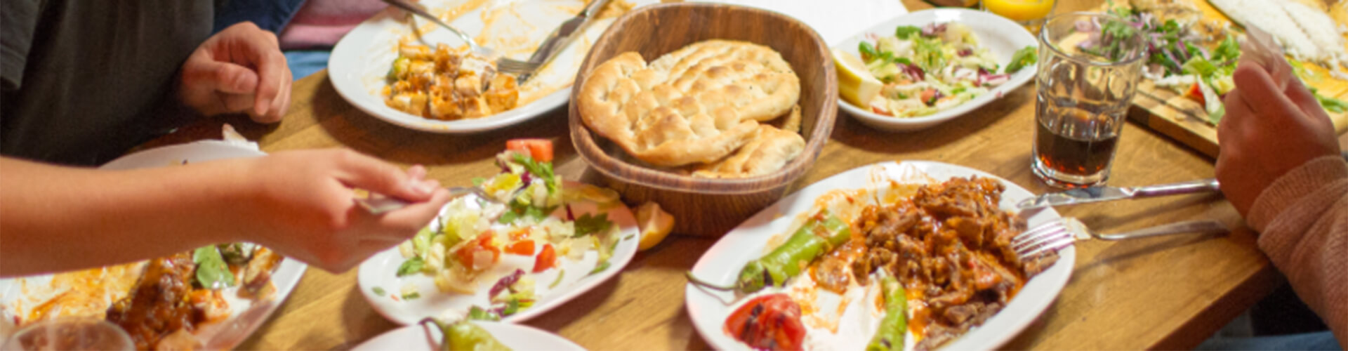 A table with various dishes including plates of food, a bowl of bread, salads, and a glass of soda. Four people are reaching for food.