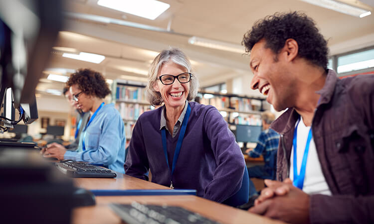 Two people sitting at computers in a library, smiling and chatting, while another person works in the background.