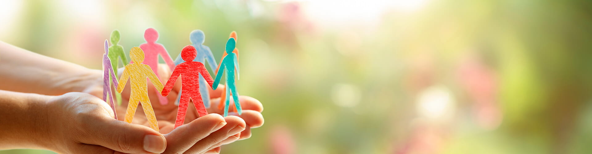 Hands holding a circle of colorful paper cutout people against a soft-focus green and pink background.