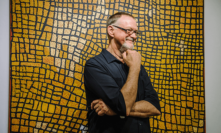 A man with glasses stands in front of an abstract yellow and black patterned First Nations artwork, smiling and resting his hand on his chin.