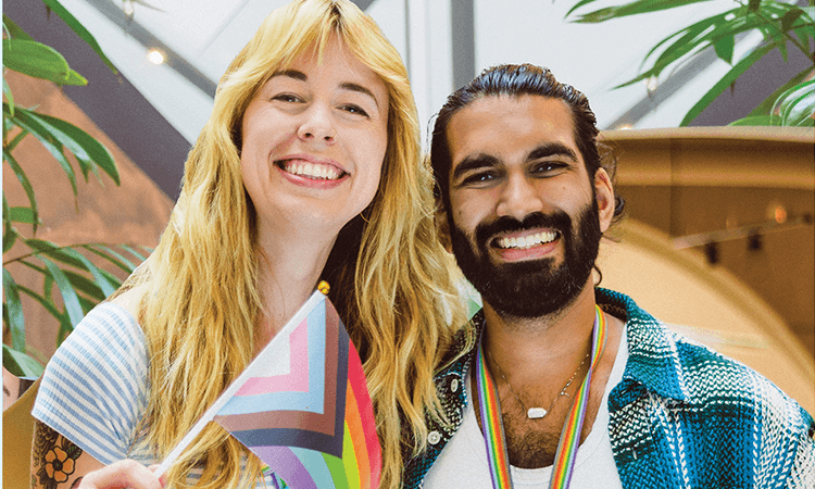 Two people smiling, one holding a Progress Pride flag, standing indoors with green plants in the background.