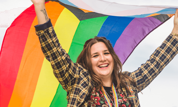 Person smiling and holding a rainbow flag with both hands, wearing a plaid jacket and a colorful lanyard, outdoors under a cloudy sky.