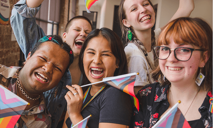 A group of five young people smile and cheer indoors, holding and waving progress pride flags.