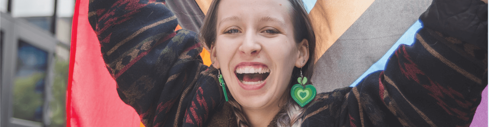 A person smiling broadly stands in front of a colorful pride flag, wearing green heart-shaped earrings and a patterned jacket.