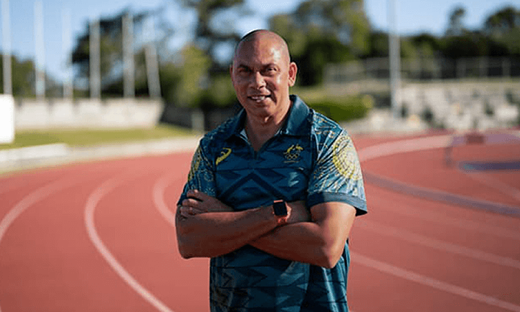 Patrick Johnson, a First Nations man, wearing a patterned sports shirt stands with arms crossed on an outdoor running track under clear skies.
