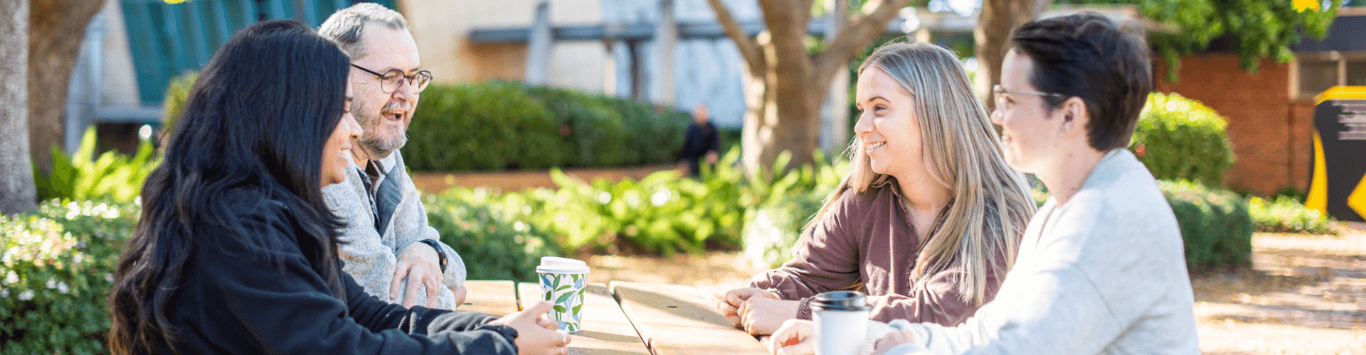 Four people sit at an outdoor picnic table, talking and holding takeaway coffee cups on a sunny day. Trees and bushes are visible in the background.