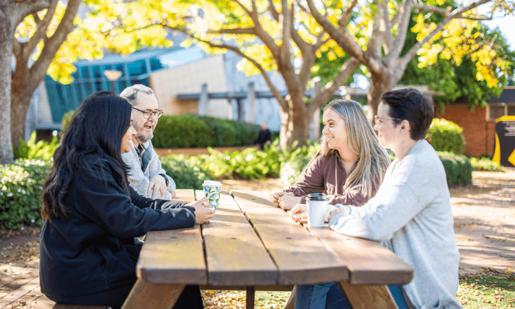 Four people sit at a wooden picnic table outdoors, talking and holding cups, with trees and buildings in the background.