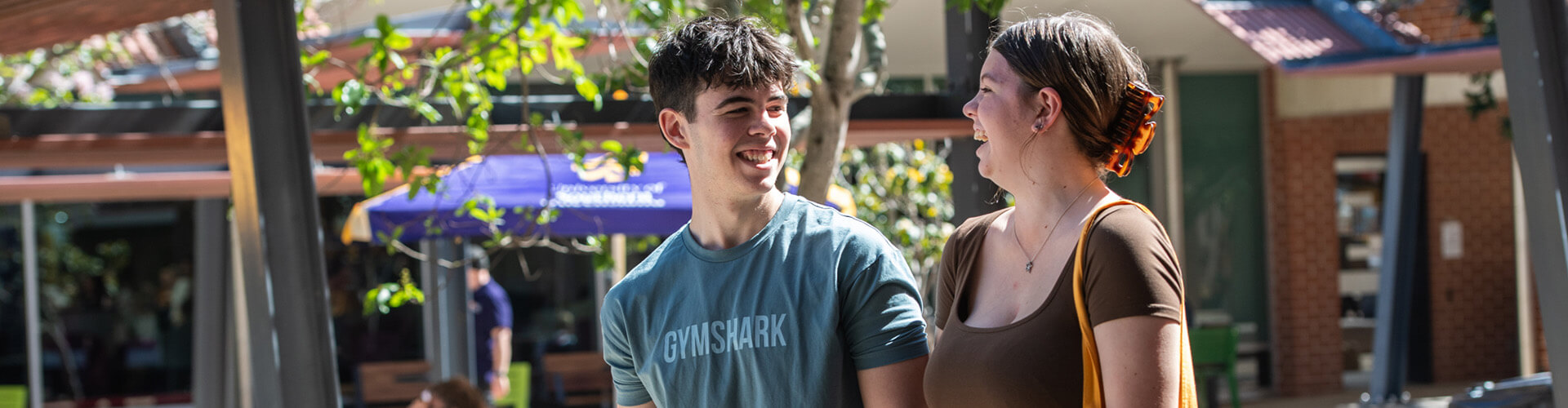 Two young adults walk outdoors on a sunny day, smiling and looking at each other, with campus buildings and trees in the background.